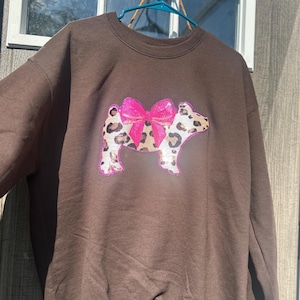 Pode incluir: Uma camisola castanha de gola redonda com um gráfico central de um porco com padrão de leopardo e um laço cor-de-rosa brilhante de lantejoulas. O design do porco é contornado por uma borda rosa, criando contraste com o tecido castanho liso.