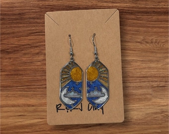 May include: A pair of dangle earrings featuring a hexagonal shape with a celestial design. The top section displays a yellow sun with glitter, while the bottom section shows a white crescent moon and clouds against a dark blue starry sky.