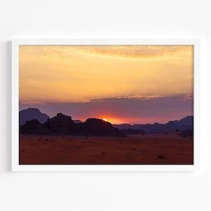 May include: A scenic landscape photograph of a desert sunset with a silhouette of rocky mountains in the foreground. The sky is a vibrant orange and purple with a bright orange sun setting behind the mountains.