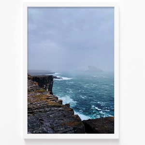 May include: A dramatic seascape photograph featuring a rocky cliff edge overlooking a stormy ocean. The waves are crashing against the shore, and the sky is overcast with clouds. A distant island is visible in the background.