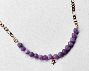 May include: A delicate necklace featuring a strand of round, lavender-colored beads. The necklace is accented with a gold-colored chain and a small star-shaped pendant with a central clear stone. The necklace is displayed on a white surface.