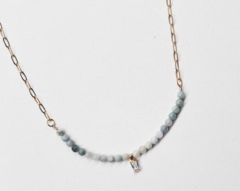 May include: A delicate gold chain necklace with a graduated ombre of light blue and gray beads. A small, clear, rectangular crystal dangles from the center of the necklace.