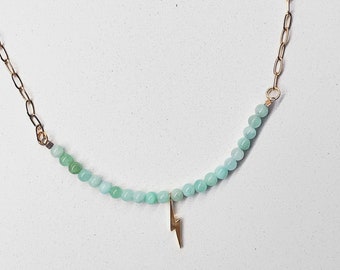 May include: A delicate gold chain necklace with a light green gemstone bead strand and a small gold lightning bolt charm.