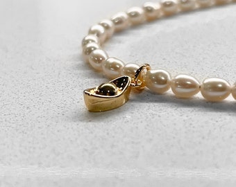May include: A white pearl bracelet with a gold charm shaped like a boat. The charm has a small round detail in the center.