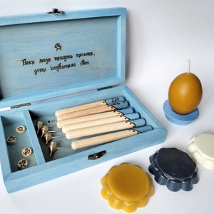 Puede incluir: Una caja de madera azul con una tapa con bisagras, que contiene un conjunto de herramientas para tallar cera. La caja tiene una cita grabada en la tapa: "Пока люди пишут писанки,  готы испытывают свет". La caja está abierta, revelando las herramientas y un pequeño compartimento con piezas de madera. También hay tres discos de cera de diferentes colores: amarillo, blanco y azul.
