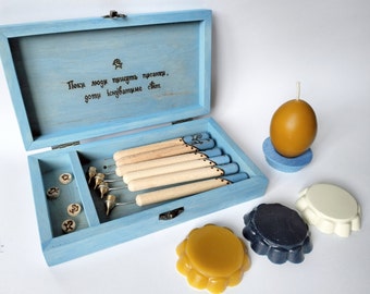 Op de afbeelding: Een blauwe houten doos met een scharnierend deksel, die een set gereedschap voor het snijden van was bevat. De doos heeft een citaat gegraveerd op het deksel: "Пока люди пишут писанки,  готы испытывают свет". De doos is open, waardoor de gereedschappen en een klein compartiment met houten stukken zichtbaar zijn. Er zijn ook drie was schijven in verschillende kleuren: geel, wit en blauw.