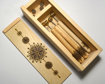 May include: A wooden box with a lid containing a set of four wooden tools with metal tips. The lid is decorated with a geometric design of snowflakes and circles. The tools are arranged in the box with their tips facing up.