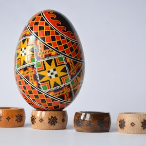 May include: A hand-painted Easter egg with geometric patterns in red, black, yellow, and green colors, resting on a wooden egg stand. The stand has a carved design of snowflakes.