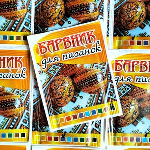 May include: Close-up of several packets of egg dye, each featuring a decorated egg and the text "БАРВНИК для писанок." The packets are orange and white, with a color palette along the bottom. The eggs are brown and orange with intricate designs.
