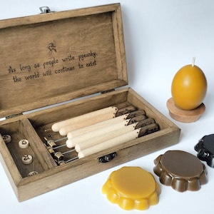 May include: A wooden box with a quote about pysanky, a traditional Ukrainian Easter egg decorating technique. The box contains tools for pysanky, including a set of wooden tools with metal tips, and a set of wax beads. The box is open, revealing the tools inside. There are also three wax eggs, one yellow, one brown, and one black, on a white surface.