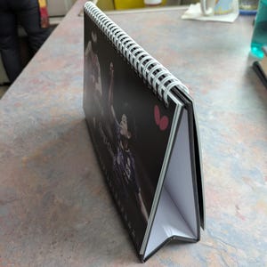 May include: A black and white desk calendar with a spiral binding. The calendar features a photo of a person playing a sport. The calendar is propped up on a table.