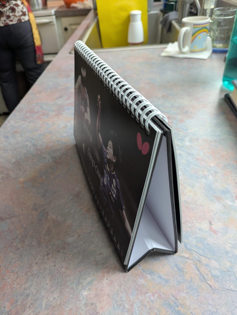 May include: A black and white desk calendar with a spiral binding. The calendar features a photo of a person playing a sport. The calendar is propped up on a table.