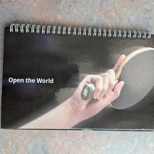 May include: A spiral-bound calendar with a black cover featuring a hand holding a ping pong paddle. The text "Open the World" is printed in white. The background is a dark gradient.