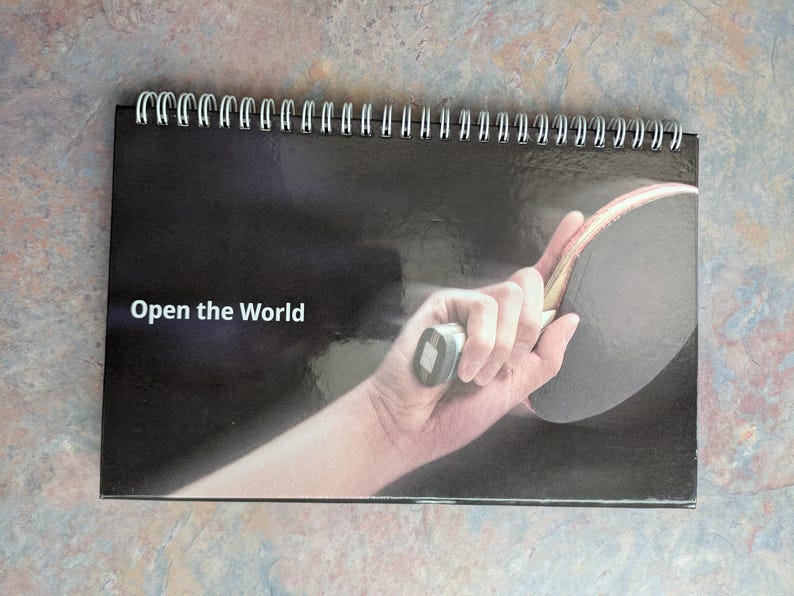 May include: A spiral-bound calendar with a black cover featuring a hand holding a ping pong paddle. The text "Open the World" is printed in white. The background is a dark gradient.
