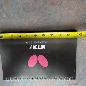 May include: A black 2026 butterfly calendar with pink butterfly wing shapes. The calendar is approximately 10 inches long, measured with a yellow tape measure. The calendar is spiral-bound.
