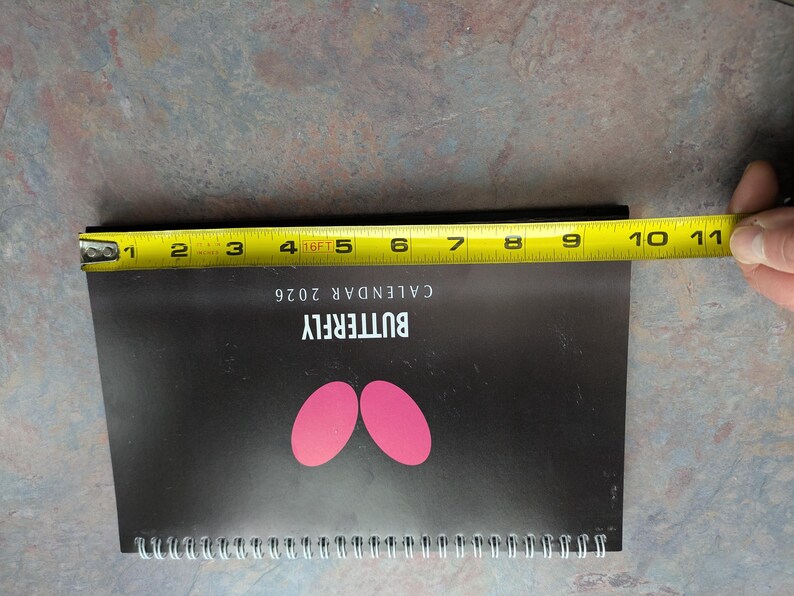 May include: A black 2026 butterfly calendar with pink butterfly wing shapes. The calendar is approximately 10 inches long, measured with a yellow tape measure. The calendar is spiral-bound.
