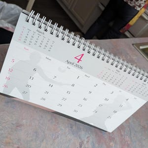 May include: A white desk calendar for April 2026, with a silver spiral binding. The calendar displays the month of April with the date of the 4th highlighted in pink. The calendar also shows the month of March and May.