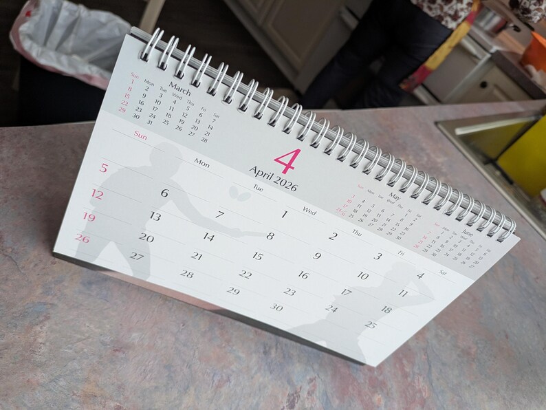 May include: A white desk calendar for April 2026, with a silver spiral binding. The calendar displays the month of April with the date of the 4th highlighted in pink. The calendar also shows the month of March and May.