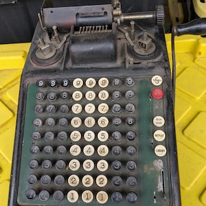 May include: An antique Burroughs Portable adding machine. The calculator has a black and green body with white and black number keys. It features a crank handle and several function buttons, including "TOTAL" and "ERROR".