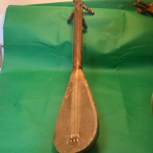 May include: A traditional stringed instrument with a long neck and a pear-shaped body. The instrument is made of wood and has a worn, aged appearance. The body is a light brown colour, and the neck is a darker brown. The instrument is resting on a green surface.
