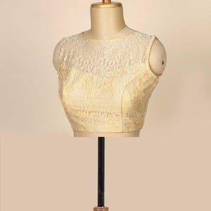 May include: A cream-coloured, sleeveless blouse on a mannequin. The blouse has a delicate floral lace overlay, giving it a textured look. The mannequin is a neutral beige, and the background is a solid, light beige.