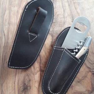 Könnte beinhalten: Zwei braune Lederscheiden mit weißen Nähten. Eine Scheide hat eine Schlaufe für einen Gürtel. Die andere Scheide hat eine Tasche für einen Flaschenöffner und einen Korkenzieher.