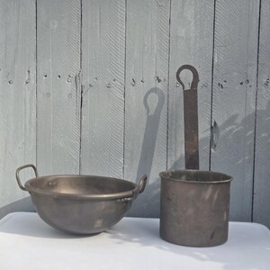 May include: A rustic, antique metal bowl with two handles and a matching cylindrical measuring cup with a long handle. The items are displayed on a white surface against a weathered gray wooden background.
