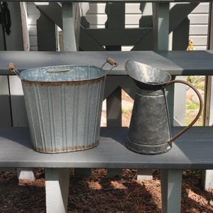 May include: Two galvanized metal containers sit on a gray bench. A ribbed bucket with wooden handles and a rusty rim is next to a pitcher with a curved handle. The items are likely for garden or home decor.