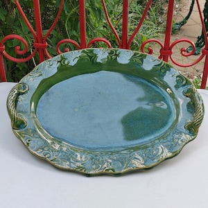 Pode incluir: Uma travessa de servir de cerâmica verde com um design floral decorativo e duas alças. A travessa tem um acabamento brilhante e uma superfície ligeiramente texturizada.