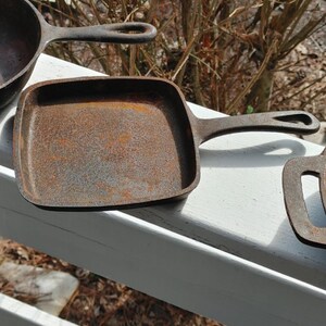 May include: Three cast iron skillets, one square and two round, with a rustic, aged, and rusty appearance. The skillets are arranged on a white wooden surface.