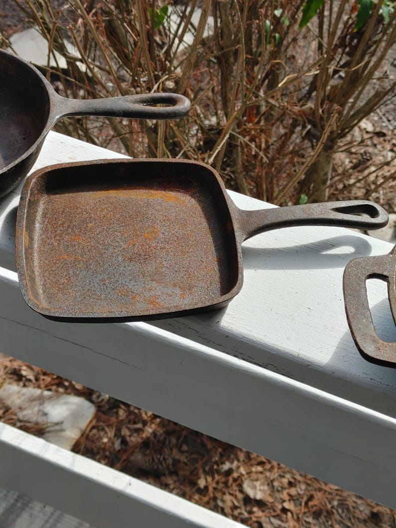 May include: Three cast iron skillets, one square and two round, with a rustic, aged, and rusty appearance. The skillets are arranged on a white wooden surface.