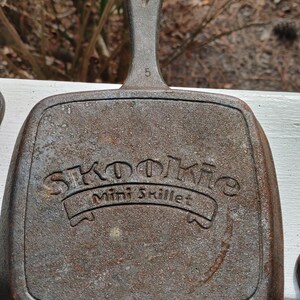 May include: A cast iron mini skillet with a brown patina. The skillet has a handle with a loop at the top and the words "Skoolie Mini Skillet" embossed on the front.