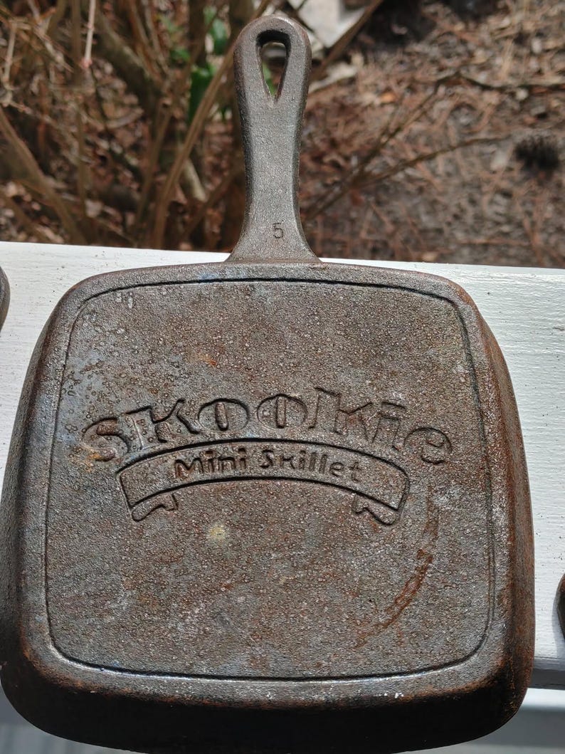 May include: A cast iron mini skillet with a brown patina. The skillet has a handle with a loop at the top and the words "Skoolie Mini Skillet" embossed on the front.