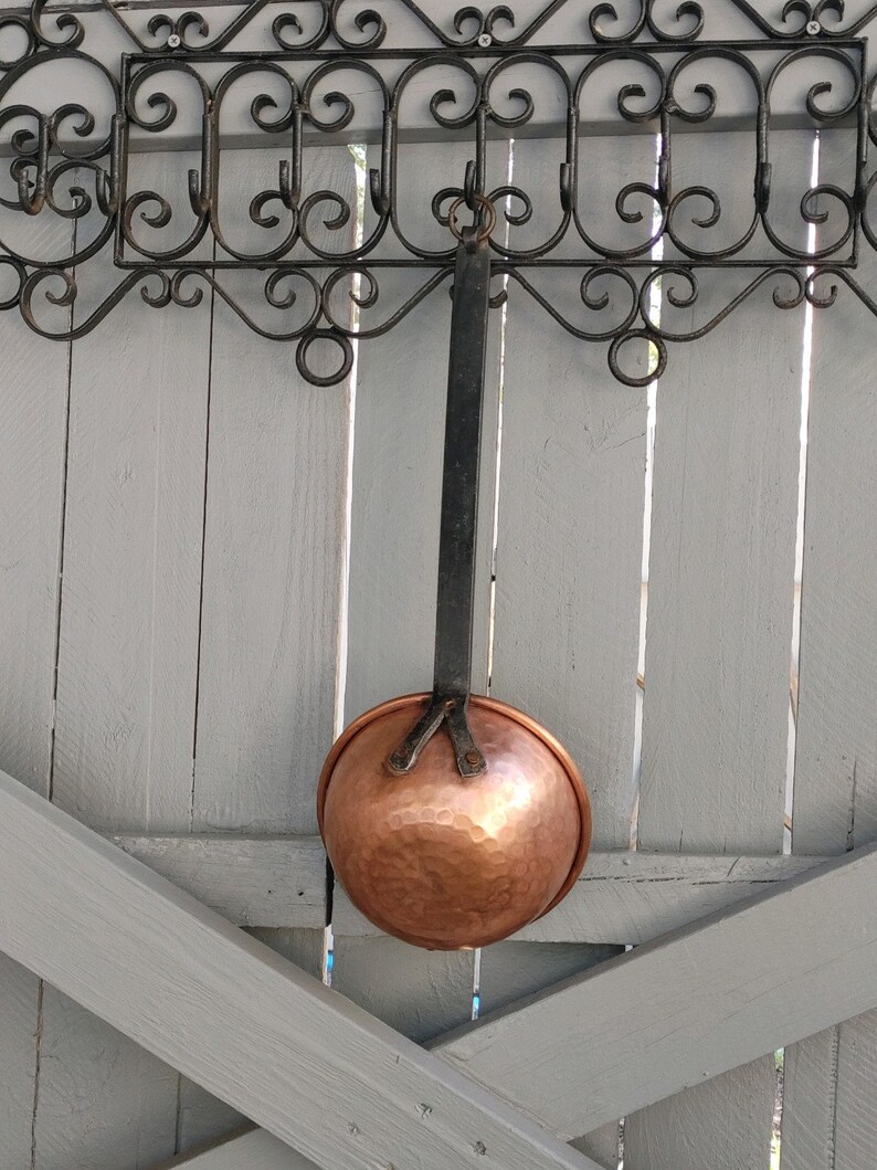 May include: A hammered copper ladle with a black leather handle hanging from a wrought iron rack.