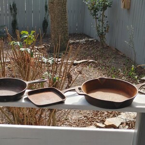 May include: Three cast iron skillets of different sizes, two with handles and one without, sitting on a white wooden surface.
