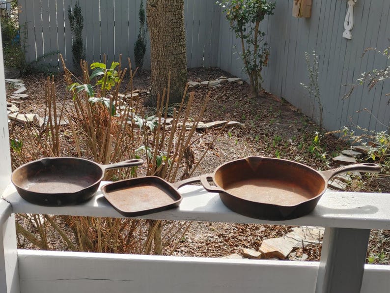 May include: Three cast iron skillets of different sizes, two with handles and one without, sitting on a white wooden surface.