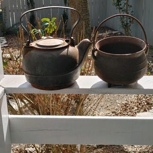 May include: Two vintage cast iron pots, one with a lid and a handle, the other with a handle, sitting on a white wooden railing.