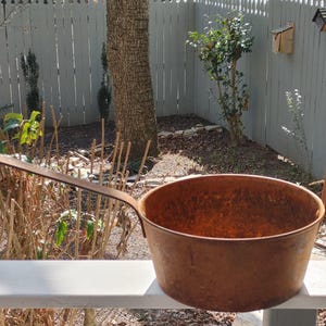 Peut inclure: Une casserole en métal rouillée avec une longue poignée et une boucle sur le côté. La casserole est posée sur une surface blanche.