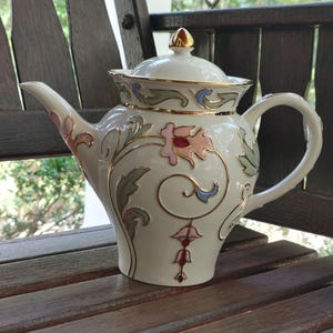 May include: A white porcelain teapot with a gold trim and floral design. The teapot has a lid with a gold knob and a spout on the side. The teapot is decorated with pink, blue, and green flowers and leaves.