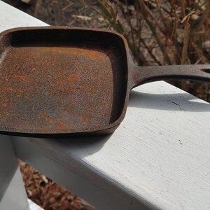 May include: A rusty cast iron square skillet with a black handle. The skillet is sitting on a white surface.