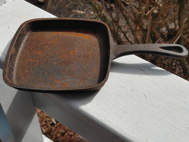 May include: A rusty cast iron square skillet with a black handle. The skillet is sitting on a white surface.