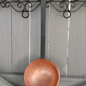 May include: A copper ladle with a hammered finish hangs from a black wrought iron rack. The ladle has a long black handle.
