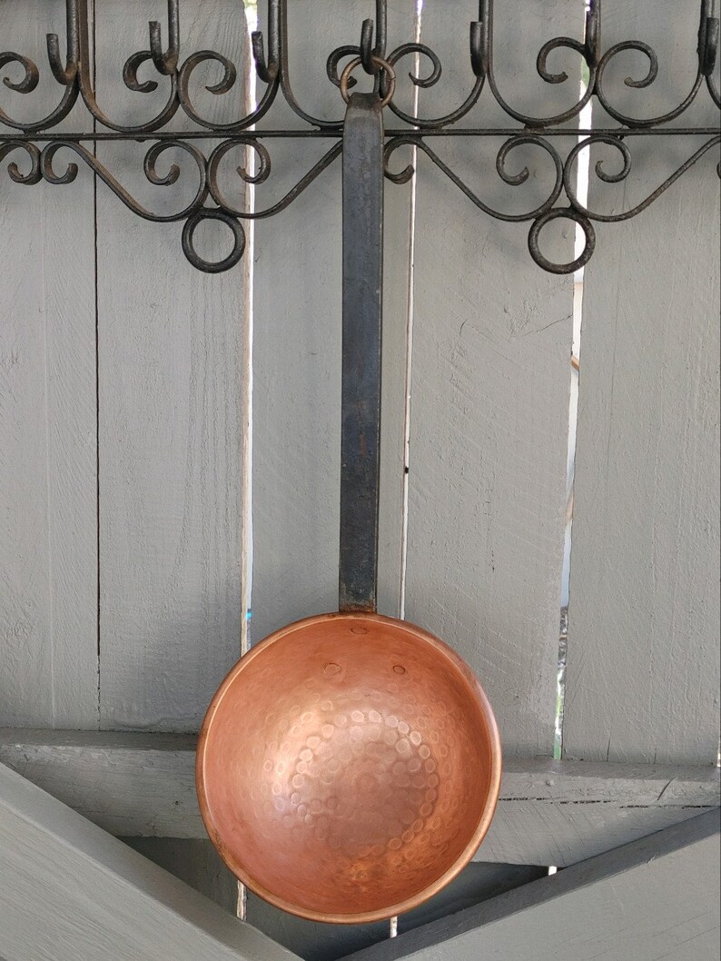 May include: A copper ladle with a hammered finish hangs from a black wrought iron rack. The ladle has a long black handle.