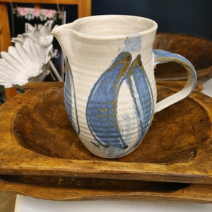 May include: A white ceramic pitcher with blue and gold abstract designs. The pitcher has a handle and is sitting on a wooden tray.