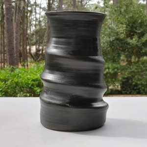 May include: A black ceramic vase with a wavy, textured design. The vase is tall and narrow, with a wide opening at the top.