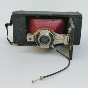 May include: A vintage wooden box camera with a red bellows and a silver lens. The camera has a black leather strap and a black cord attached to the lens.