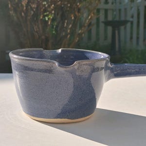 May include: A blue ceramic serving bowl with a handle. The bowl has a speckled glaze and a slightly rounded shape.