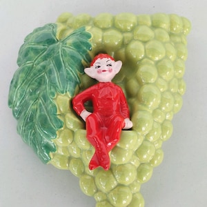 May include: A ceramic wall hanging depicting a green grapevine with a red elf figurine perched on top. The elf has a playful expression and is wearing a red suit.