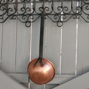 May include: A copper ladle hanging from a wrought iron rack against a light gray wooden wall.