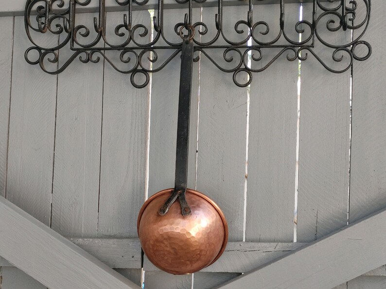 May include: A copper ladle hanging from a wrought iron rack against a light gray wooden wall.